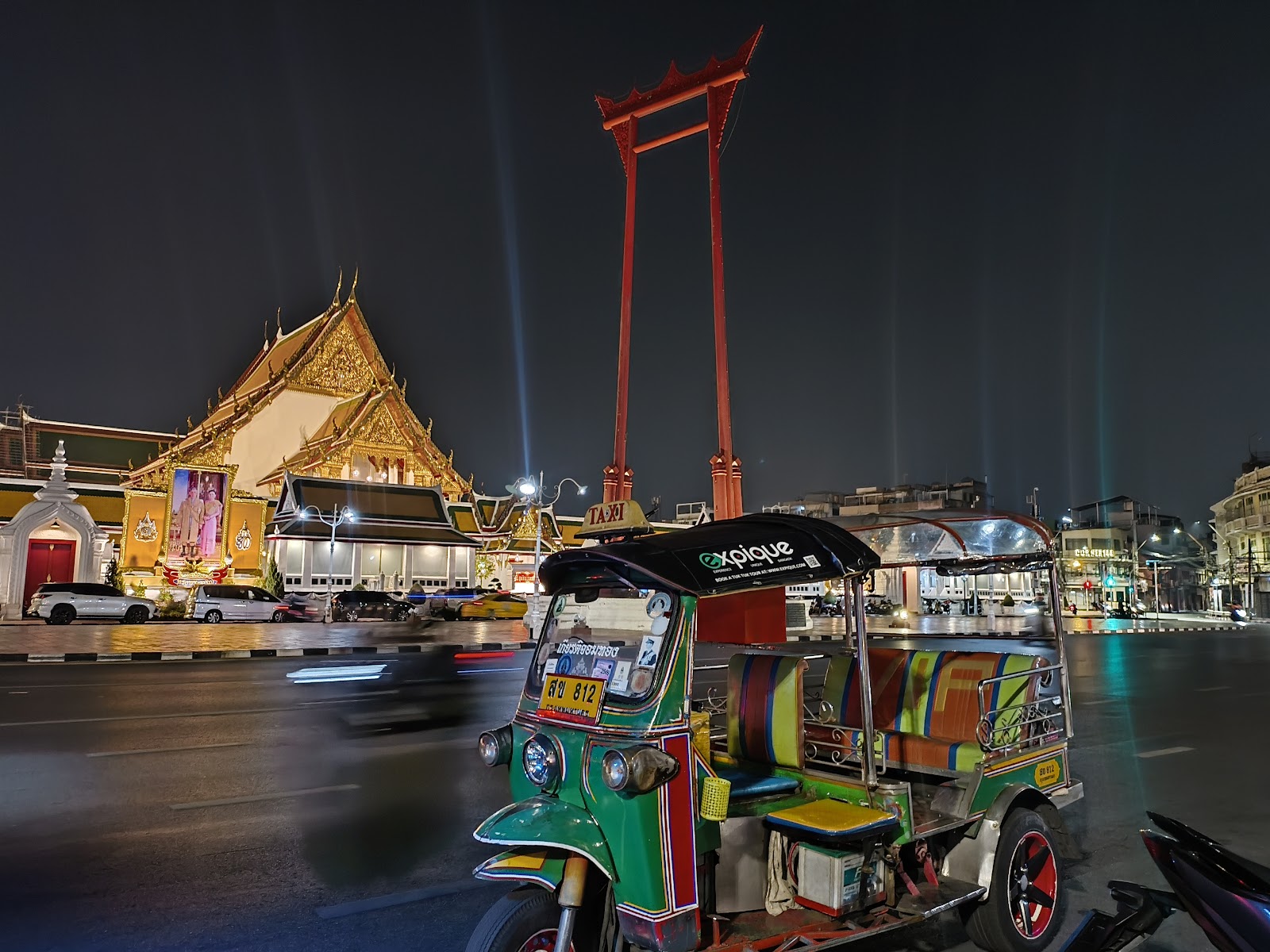 The red Giant Swing lit at night beside a temple, with a tuk-tuk in the foreground