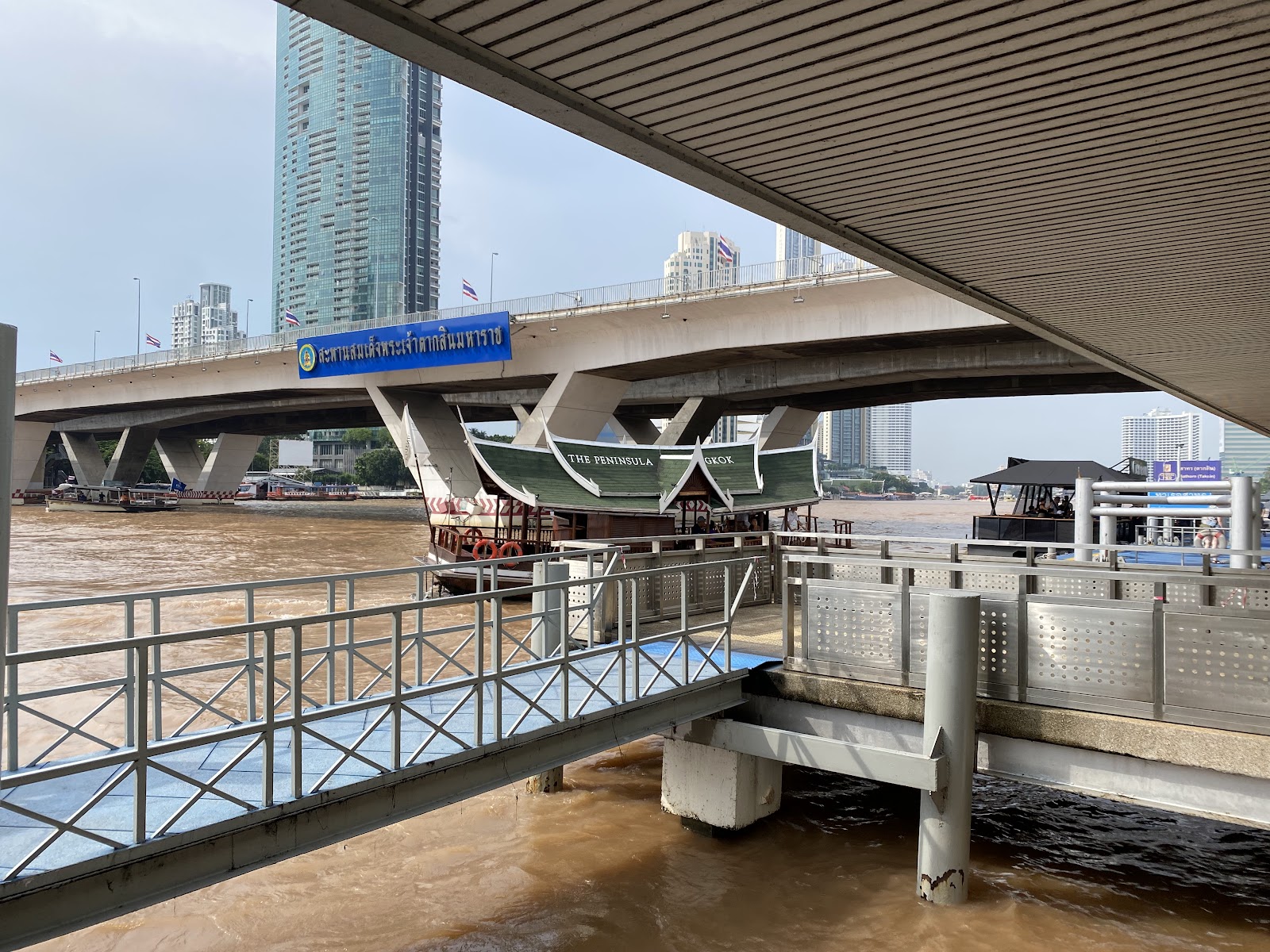 The floating walkway at Sathorn Pier on the Chao Phraya River, with the Taksin Bridge overhead and a riverside tower beyond