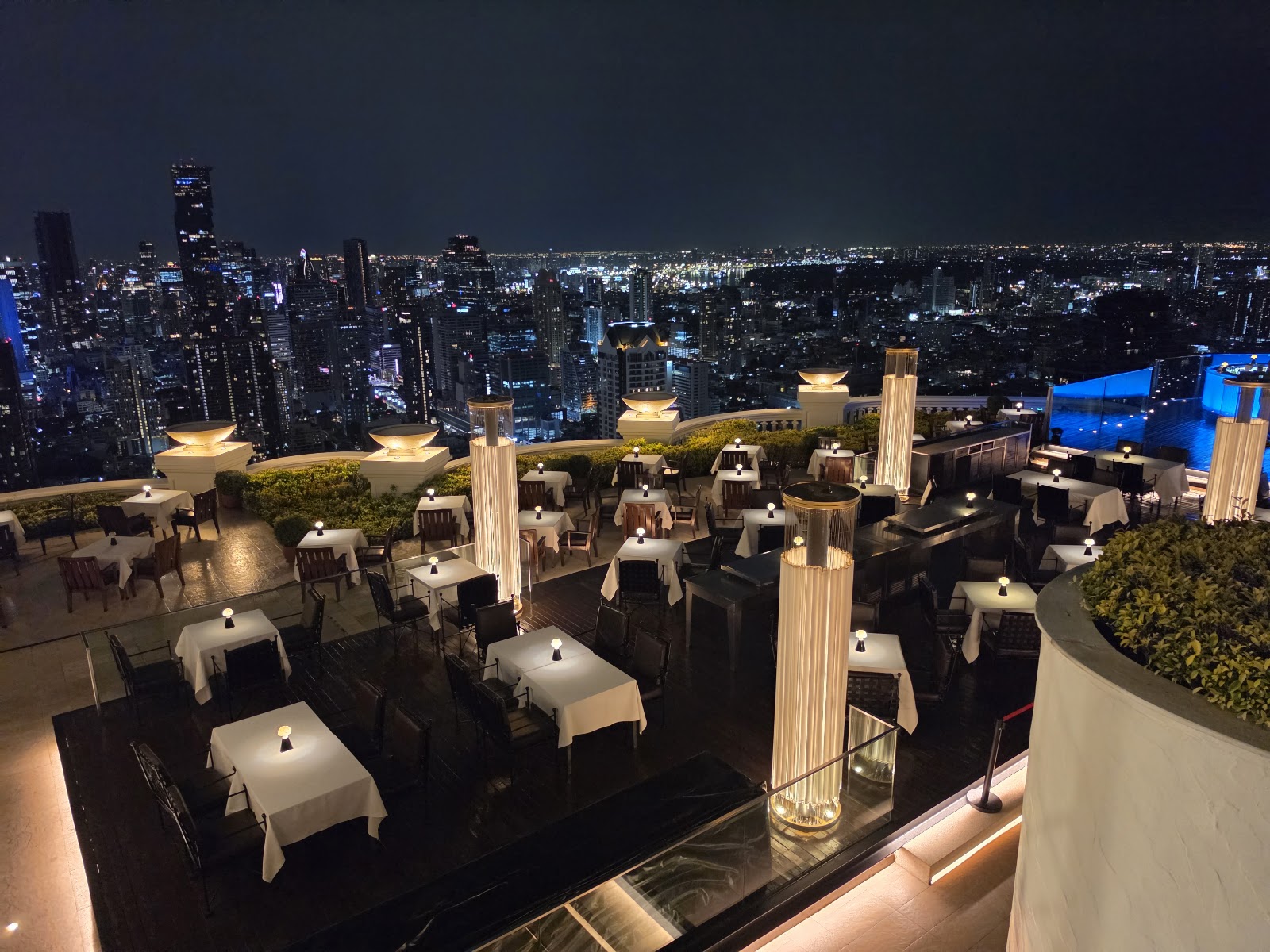 Tables on the open-air terrace of Sirocco at State Tower at night, with the lit Bangkok skyline behind