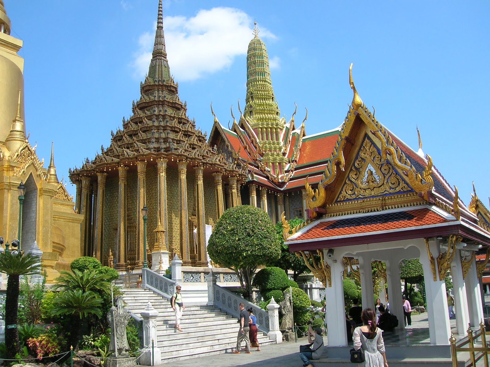 A tiered-roof hall and golden chedi inside the Grand Palace grounds under a blue sky