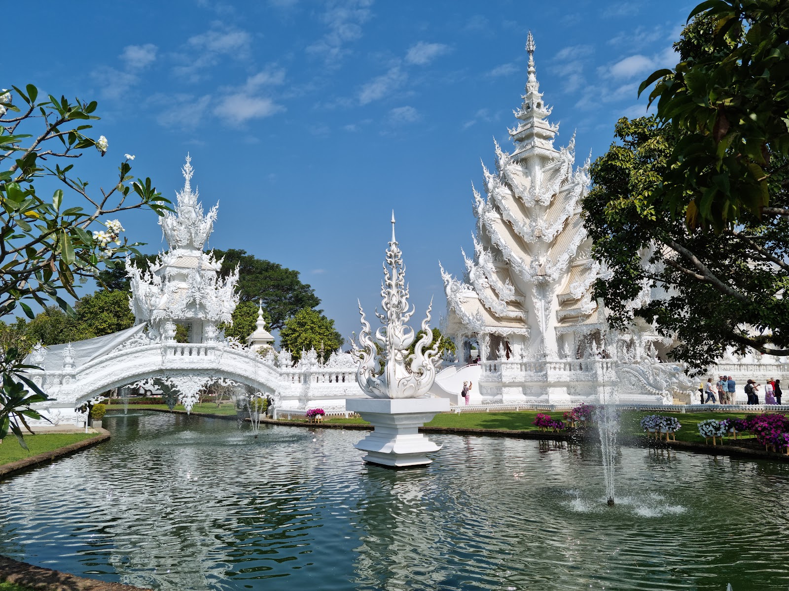 The white, ornately carved buildings of Wat Rong Khun reflected in a pond with a fountain