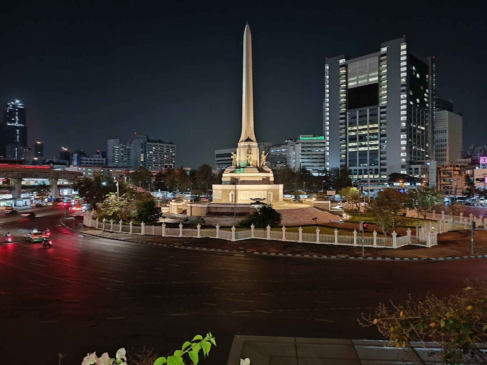 The lit obelisk of the Victory Monument in its roundabout at night, with office towers behind