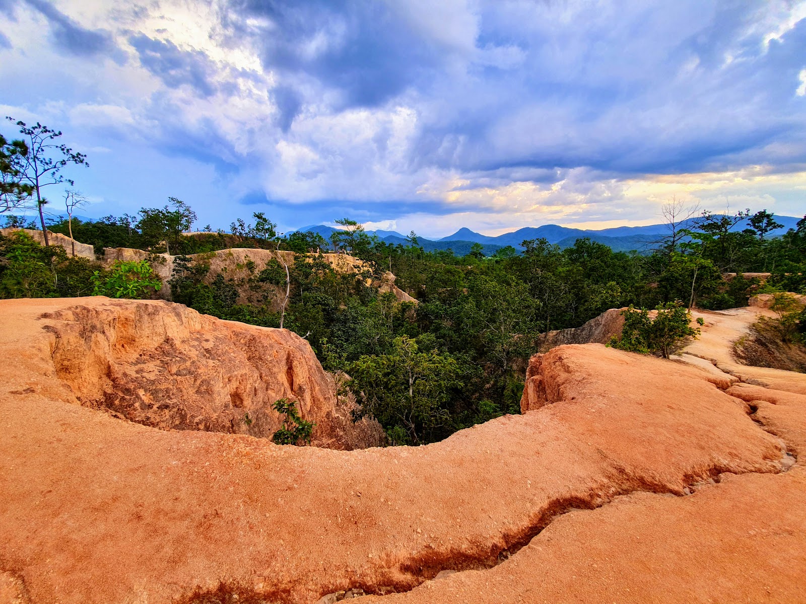Eroded orange ridges of Pai Canyon with forest and mountains behind under a cloudy sky