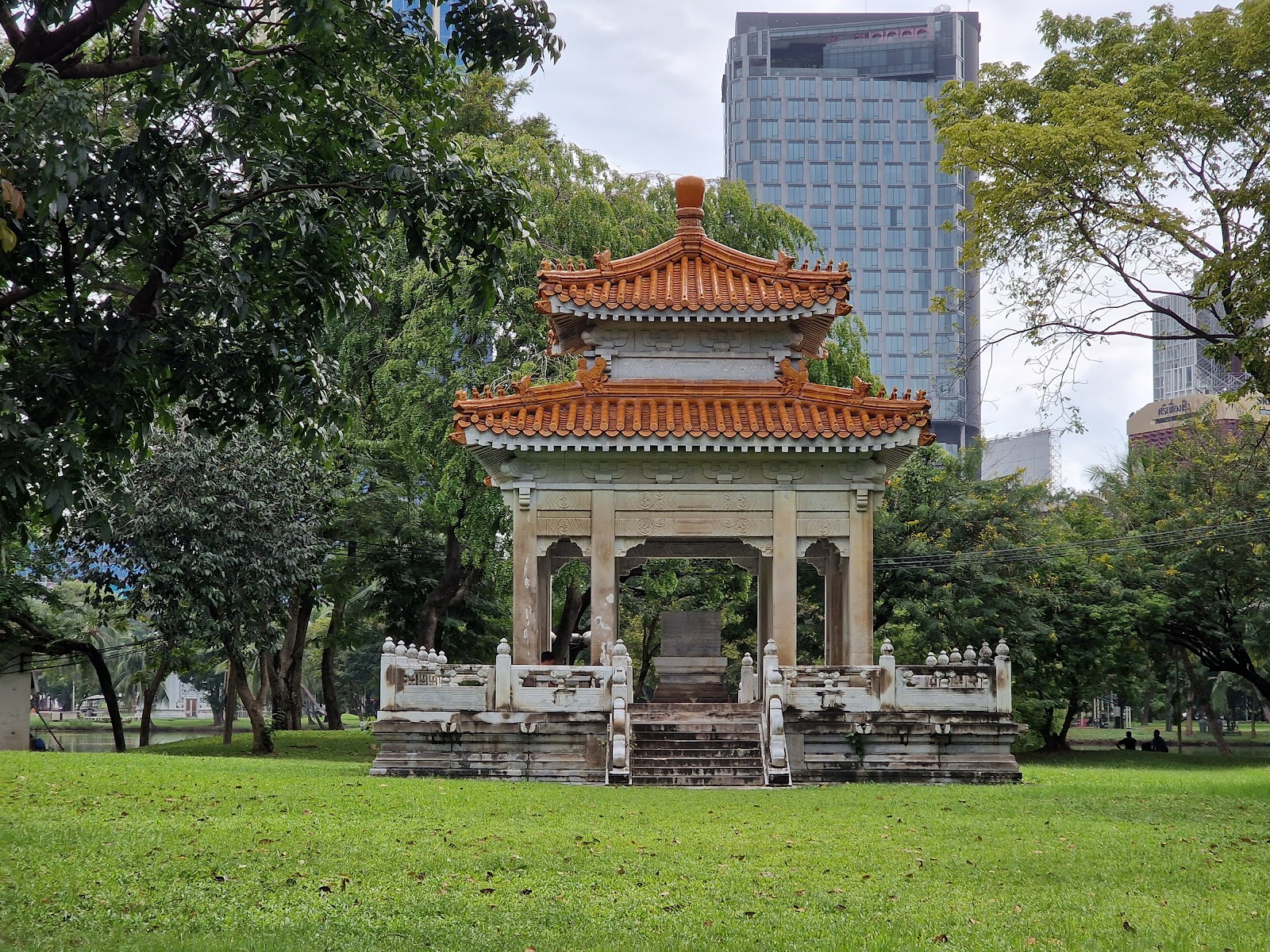 A red and white Chinese-style pavilion on a lawn in Lumphini Park with an office tower behind