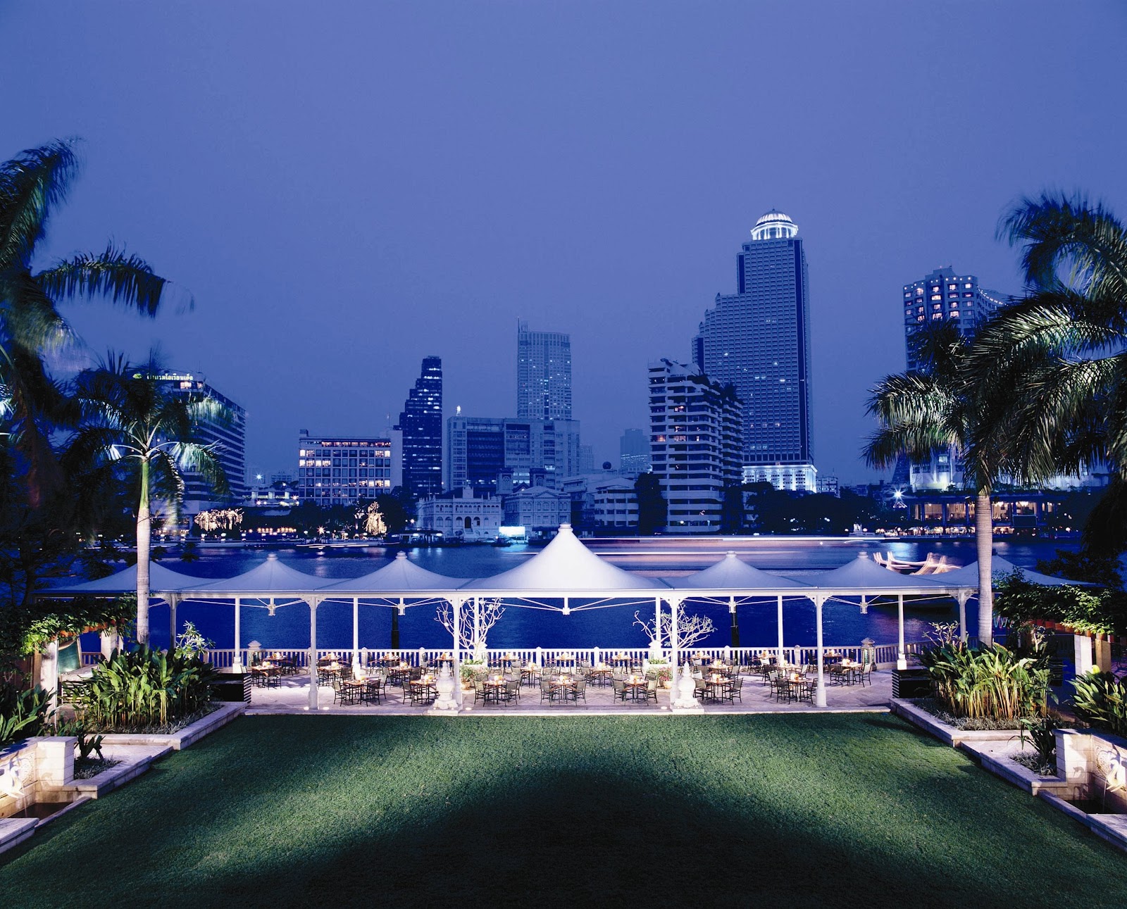 The Peninsula Bangkok's riverside lawn and white pavilions at dusk, with the city skyline across the Chao Phraya