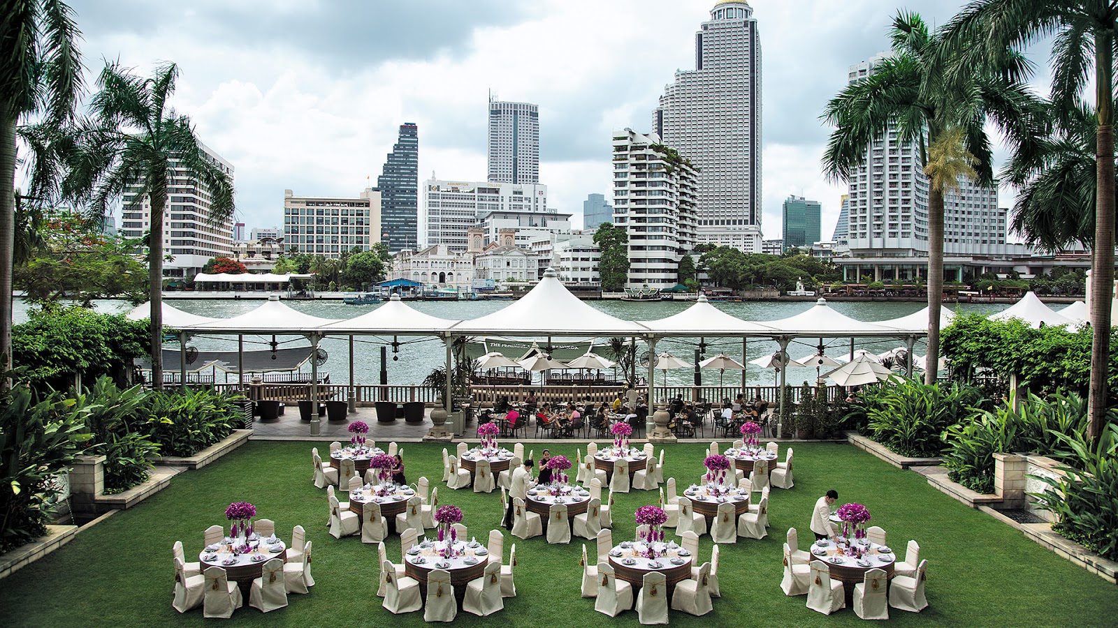 Tables set on a lawn by the river with high-rise towers across the water