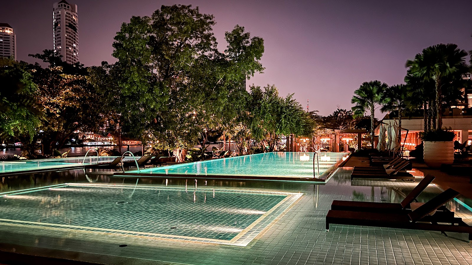 An infinity pool lit at night among trees beside the river