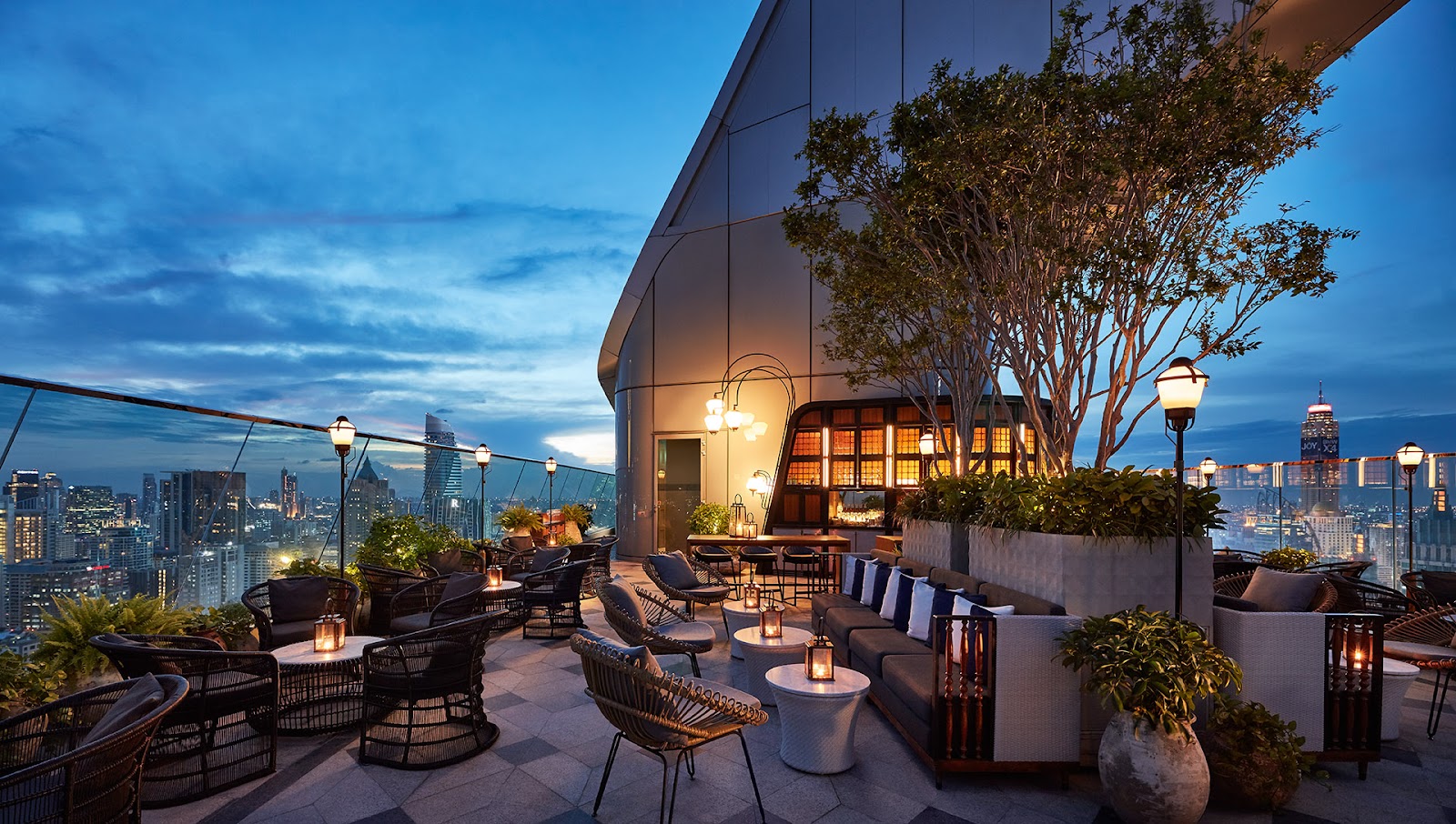 Open-air rooftop terrace with lounge seating and trees above the Bangkok skyline at twilight