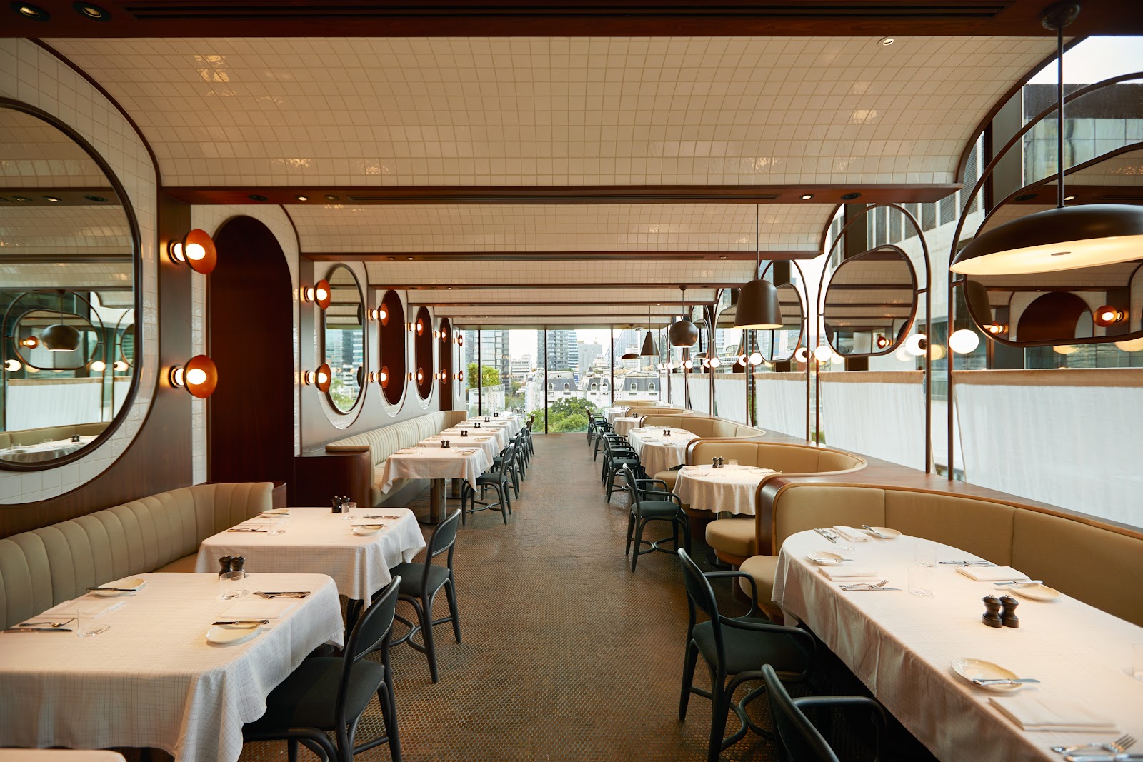 Long restaurant with white tablecloths, curved banquettes, round mirrors and a wall of windows