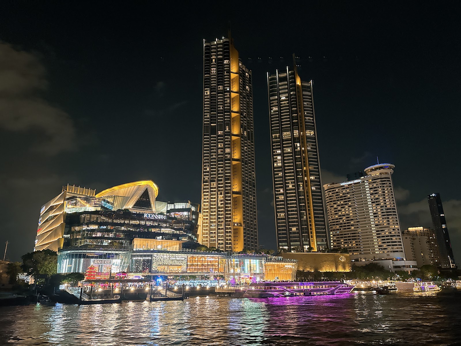 ICONSIAM and its twin residential towers lit up at night above the Chao Phraya, with boats at the pier