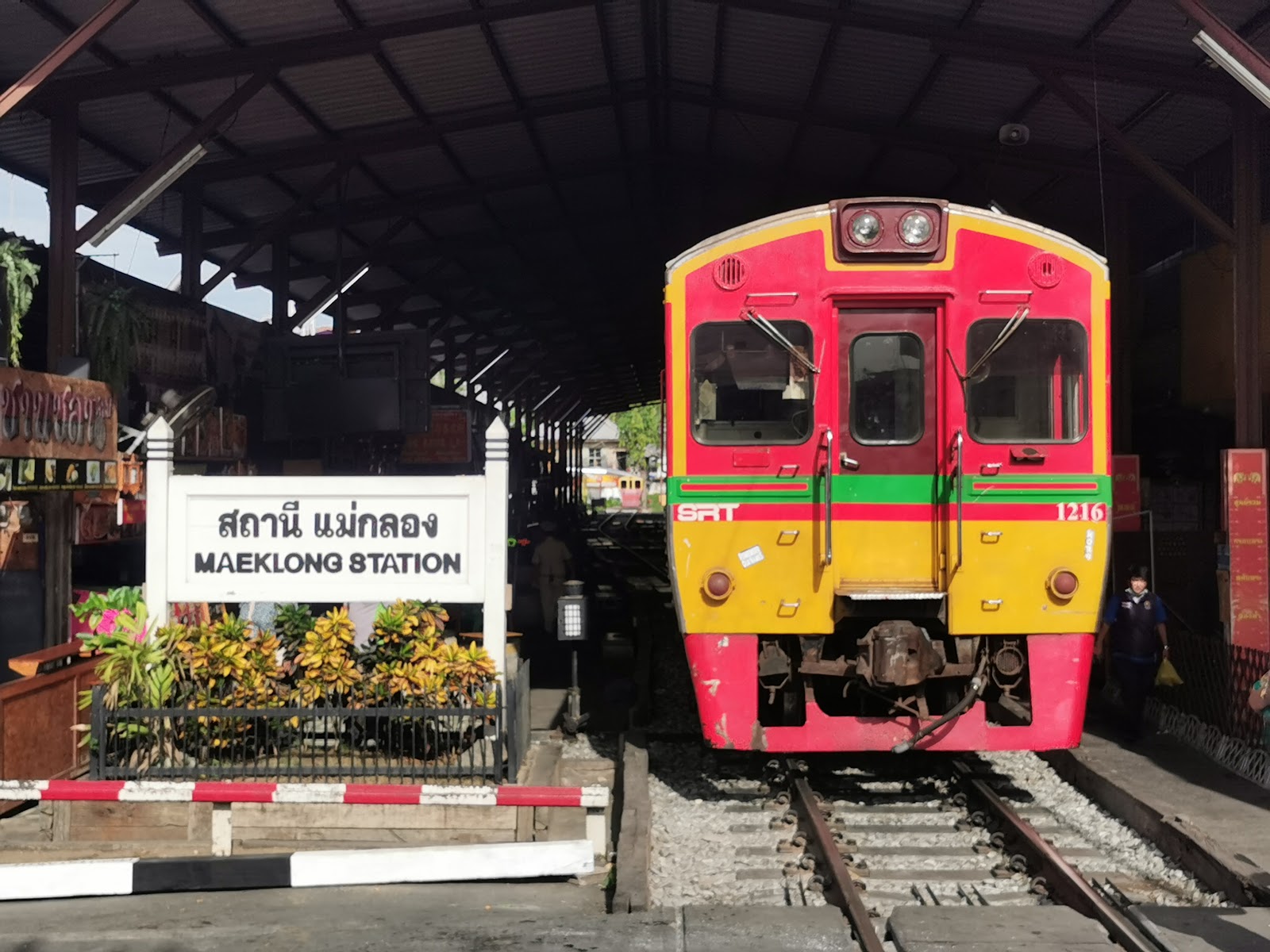 A red and yellow train standing beside the Maeklong Station sign