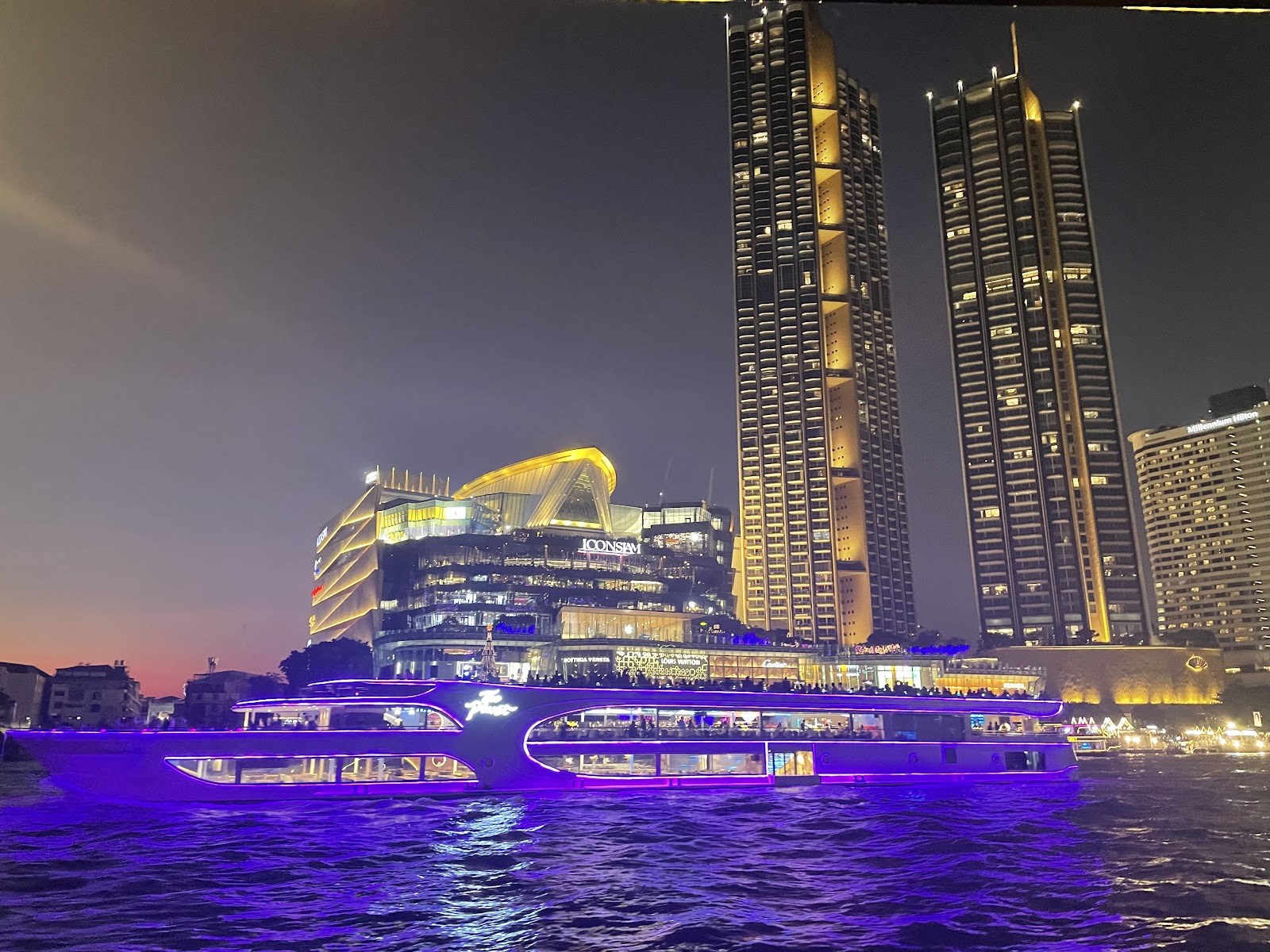 A large dinner cruise boat lit up in purple on the Chao Phraya River at night beside two tall riverside towers