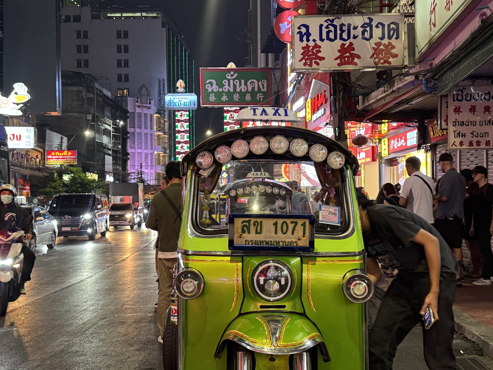A green tuk-tuk parked on a street at night near Khao San Road with shop signs lit behind it