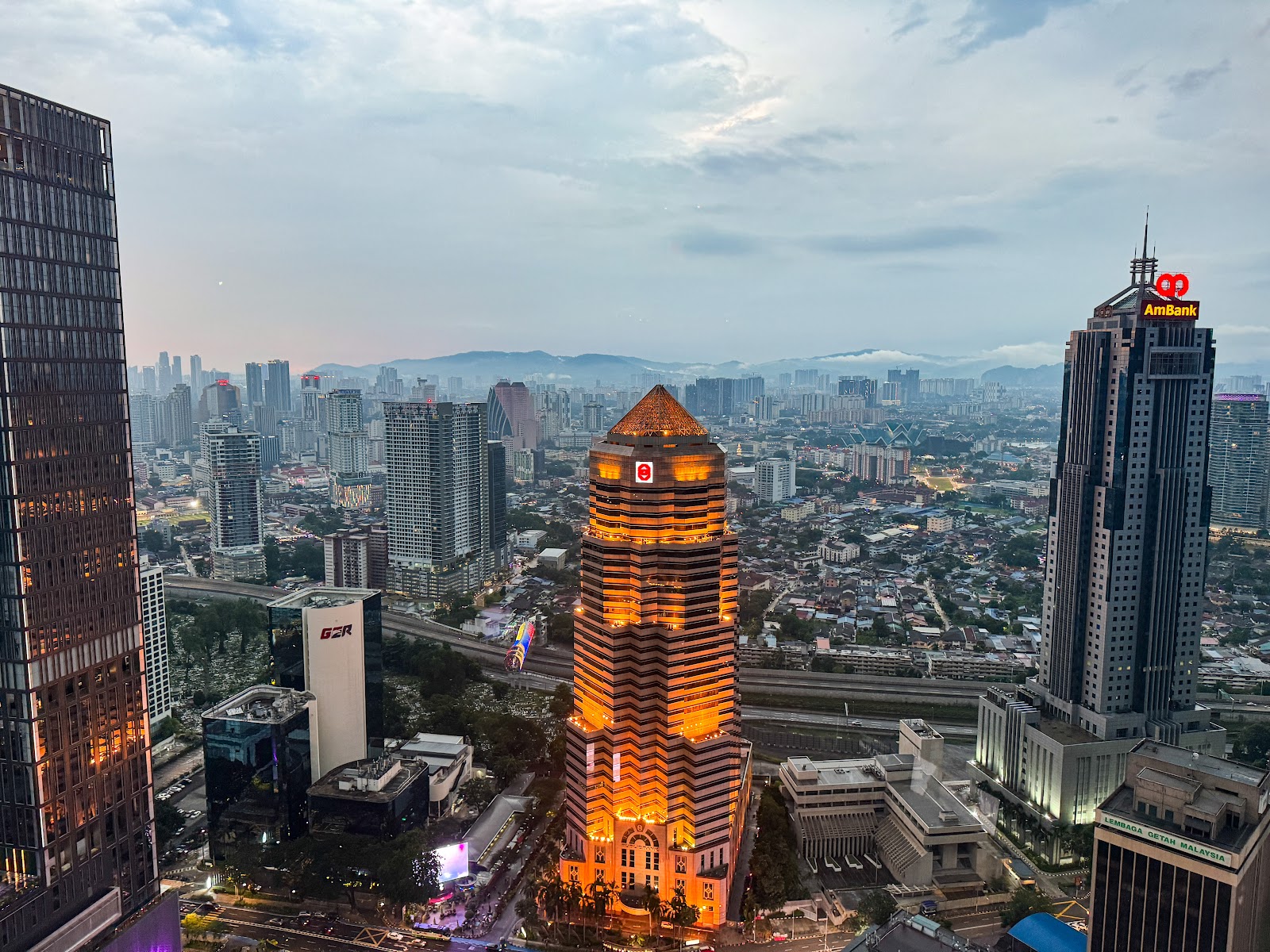 Central Kuala Lumpur seen from above, with tower blocks, a lit heritage-style building and hills on the horizon