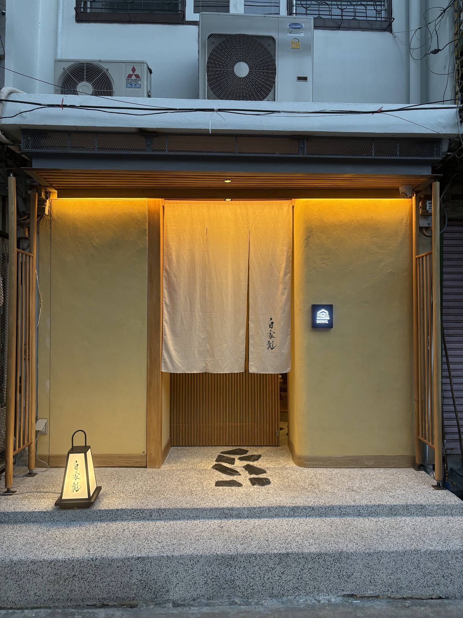 The entrance of Jikasei B, with a cream noren curtain and a small paper lantern on the step