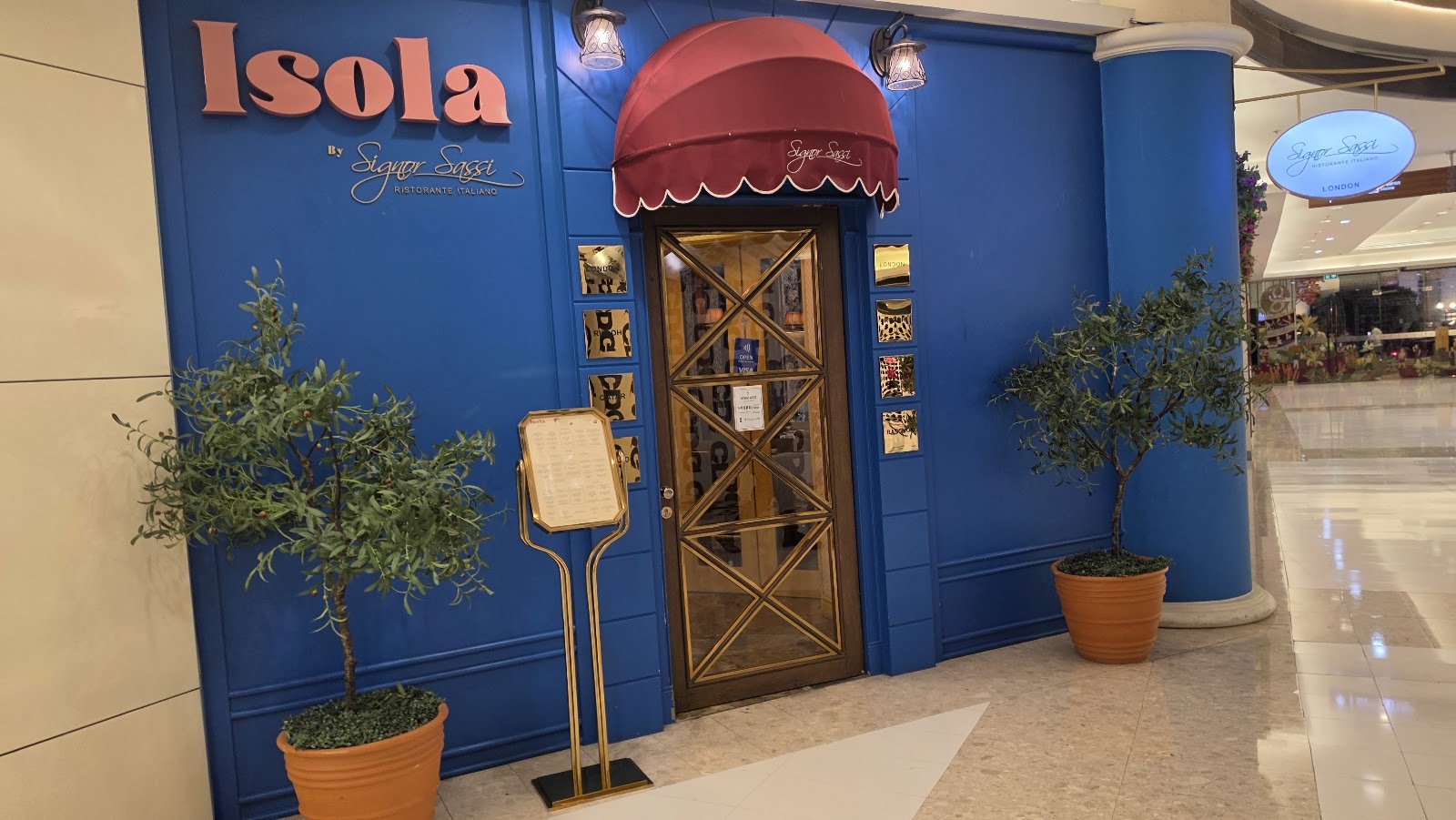 The blue entrance of Isola with a red awning and potted olive trees inside the mall