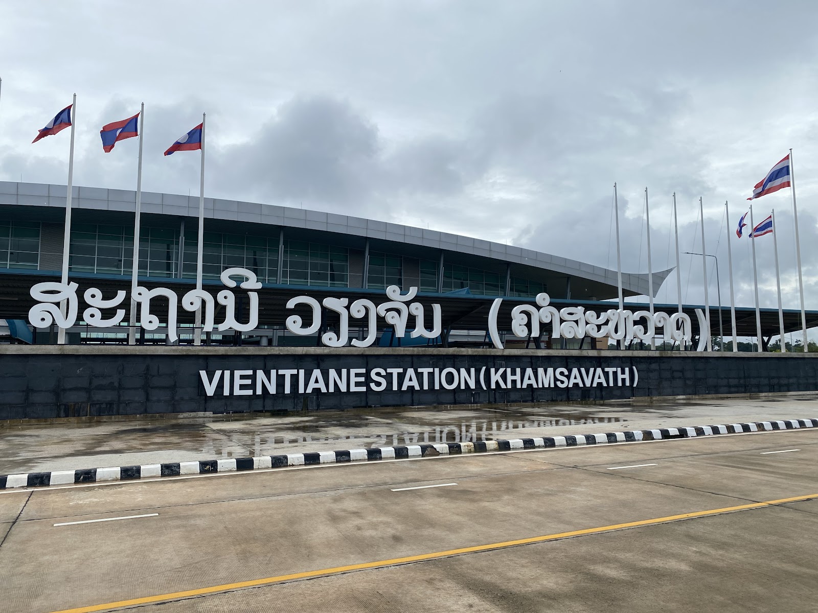 Large Lao and English lettering reading Vientiane Station (Khamsavath) in front of the modern station building with flags