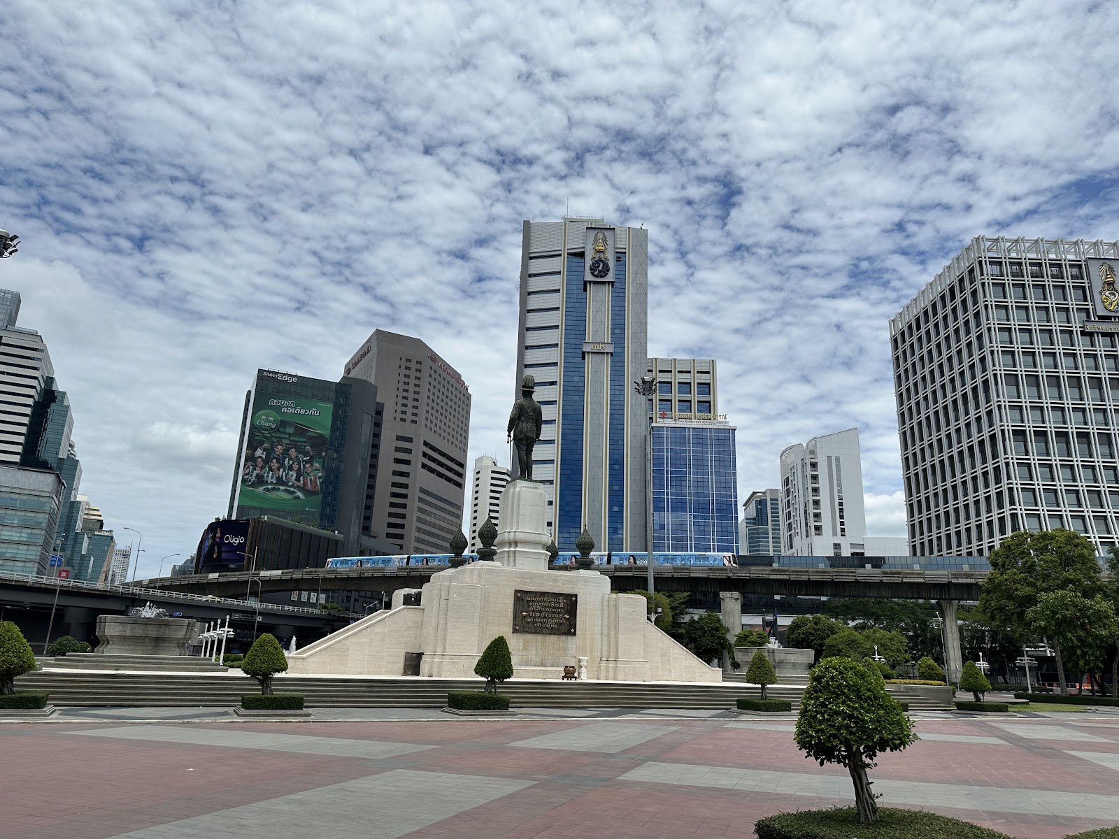 The King Rama VI statue at Lumphini Park with office towers and an elevated rail line behind it