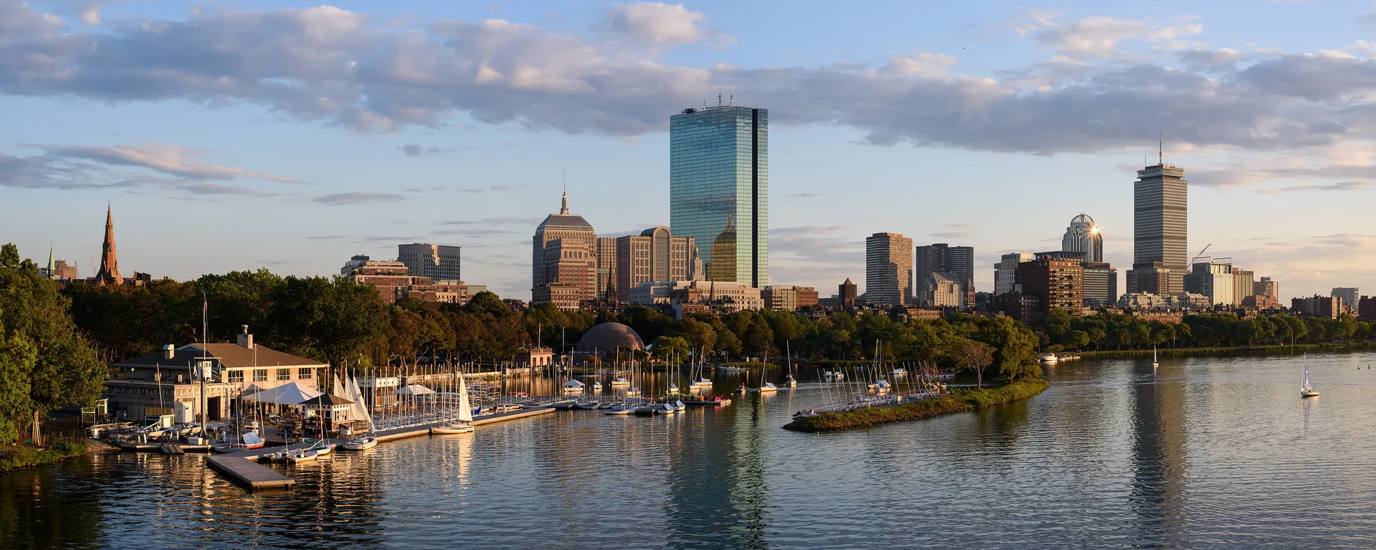 Boston skyline from the Longfellow Bridge at golden hour, with sailboats on the Charles River and the Hancock and Prudential towers